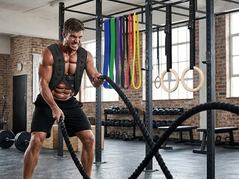 Athlete using battle ropes beside a functional training rack with resistance bands and wooden gymnastic rings.